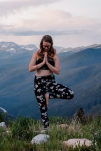 female standing on top of mountain doing a yoga tree pose