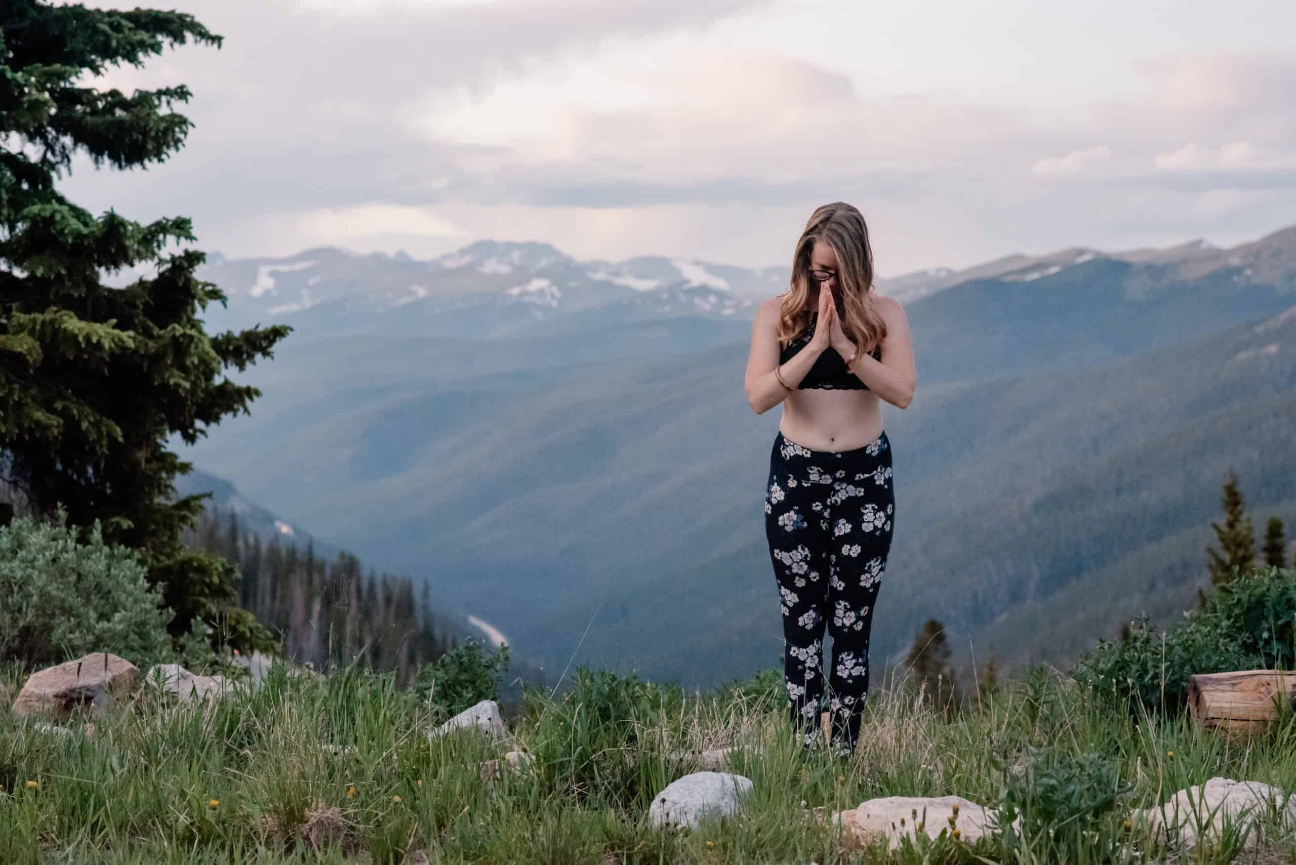 emmy bowing in self love yoga pose of namaste hands on top of a mountain after sunset