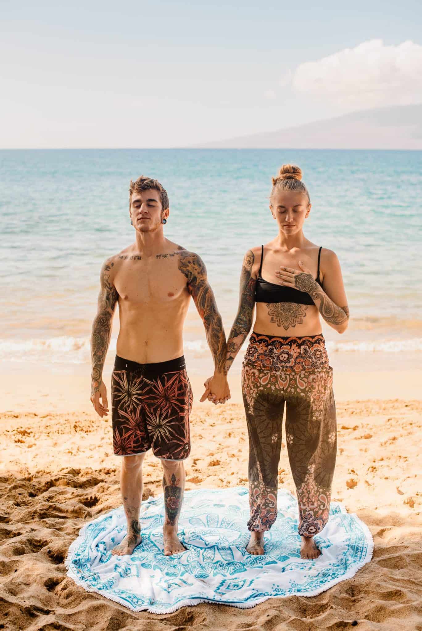 couple holding hands doing yoga for self care as female places hand on her heart while they are on the beach