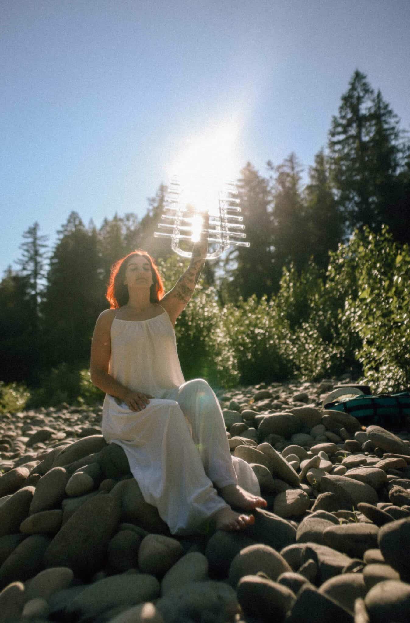 Taylor, sound bath practioner, sitting on a rock holding a chime up the sky